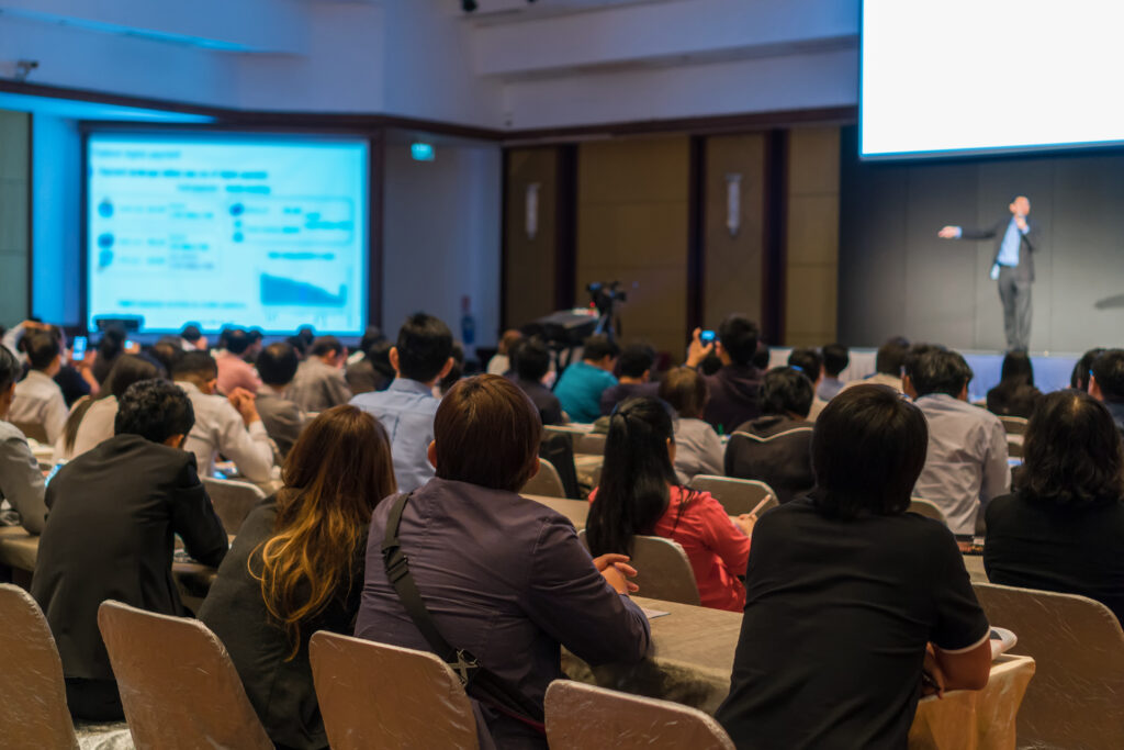 Rear side of Audiences sitting and listening the speackers on the stage in low light conference hall, event and seminar concept